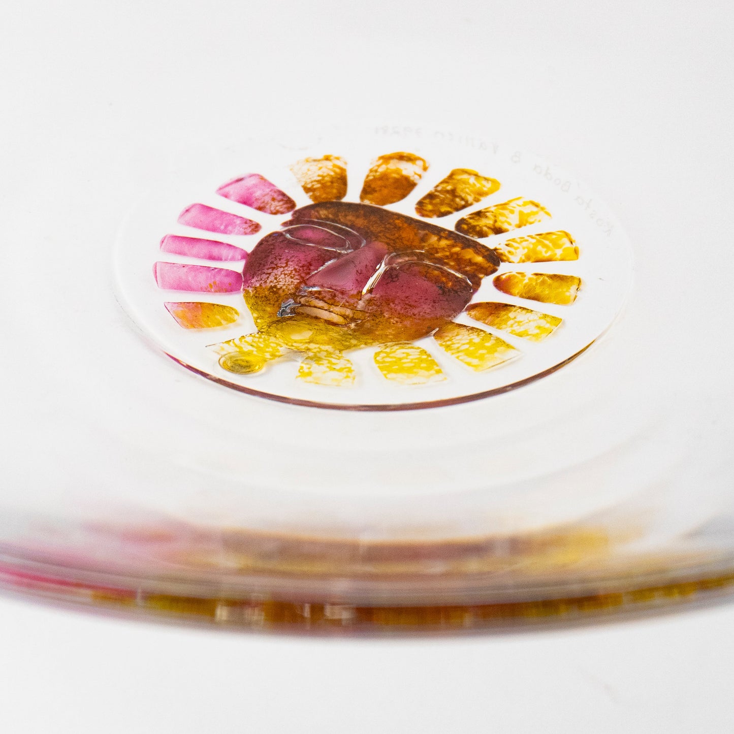 A close-up of a clear circular glass dish or plate resting on a brushed silver metal surface, with a small colorful floral sun inset containing pink, yellow, and orange details.