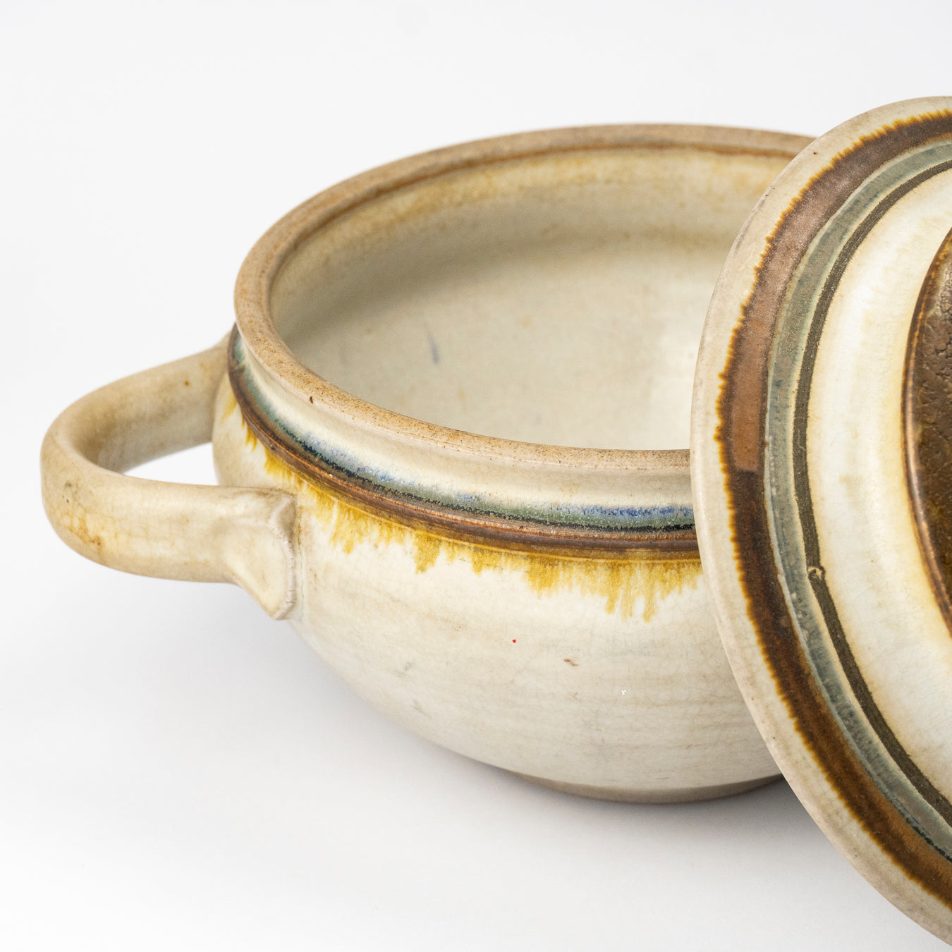 A handmade ceramic lidded pot with a single loop handle, featuring a cream-colored glaze with brown and muted blue-green accents, shown on a white background.