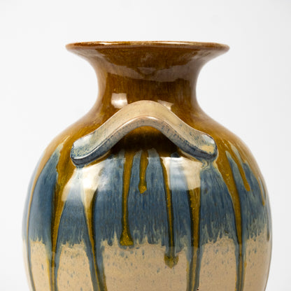 Close-up of a ceramic vase with a flared rim, a curved side handle, and a glossy glaze featuring blue, brown, and beige drip patterns on a white background.