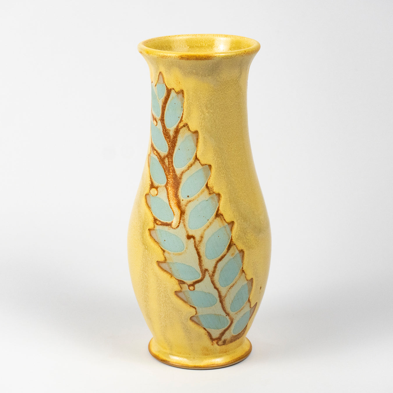 A yellow ceramic vase with a vertical leaf pattern in light blue and brown glaze, photographed on a white studio background.