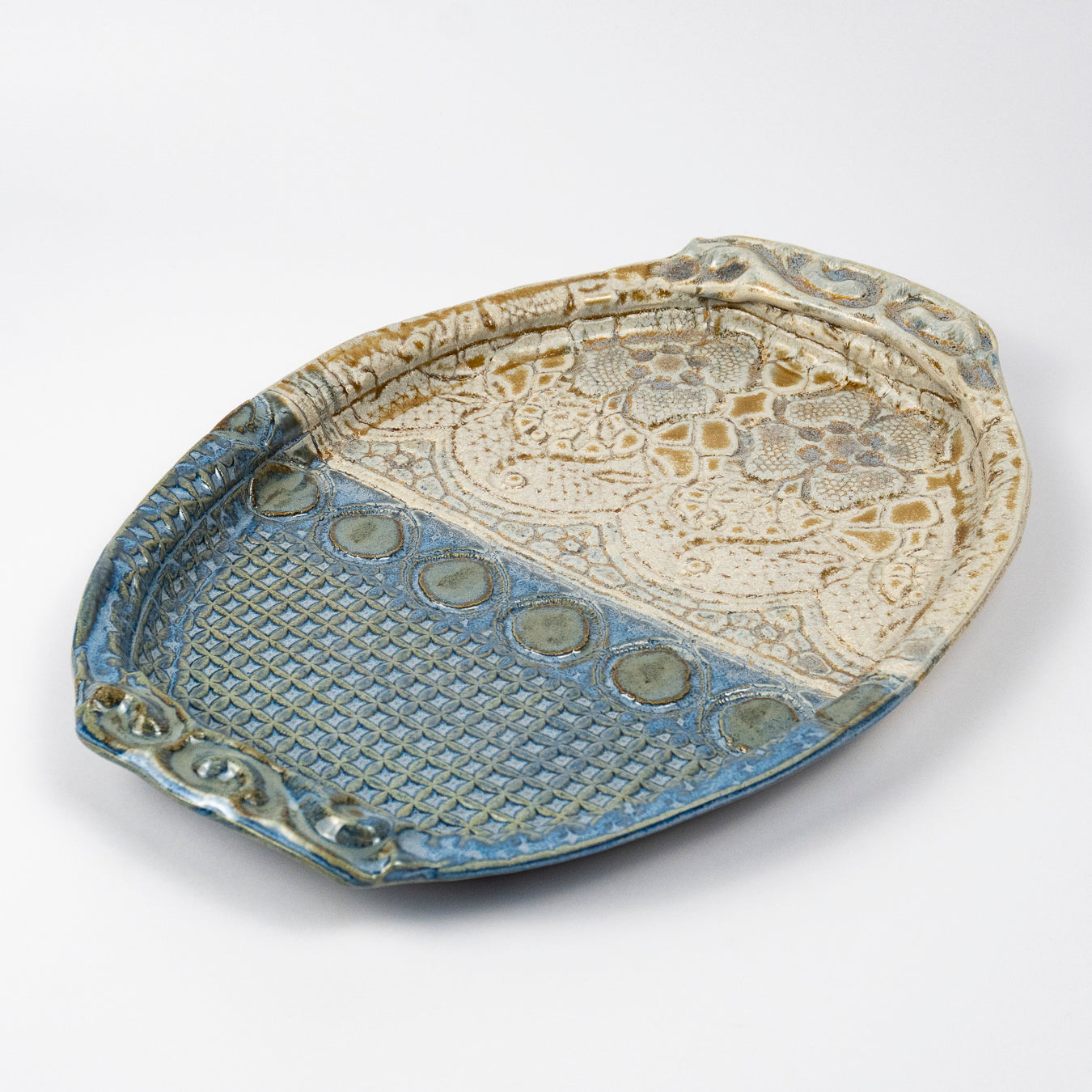 An oval ceramic serving tray with a split design, featuring a textured blue glazed lower half and an embossed cream floral patterned upper half, set against a plain light gray background.