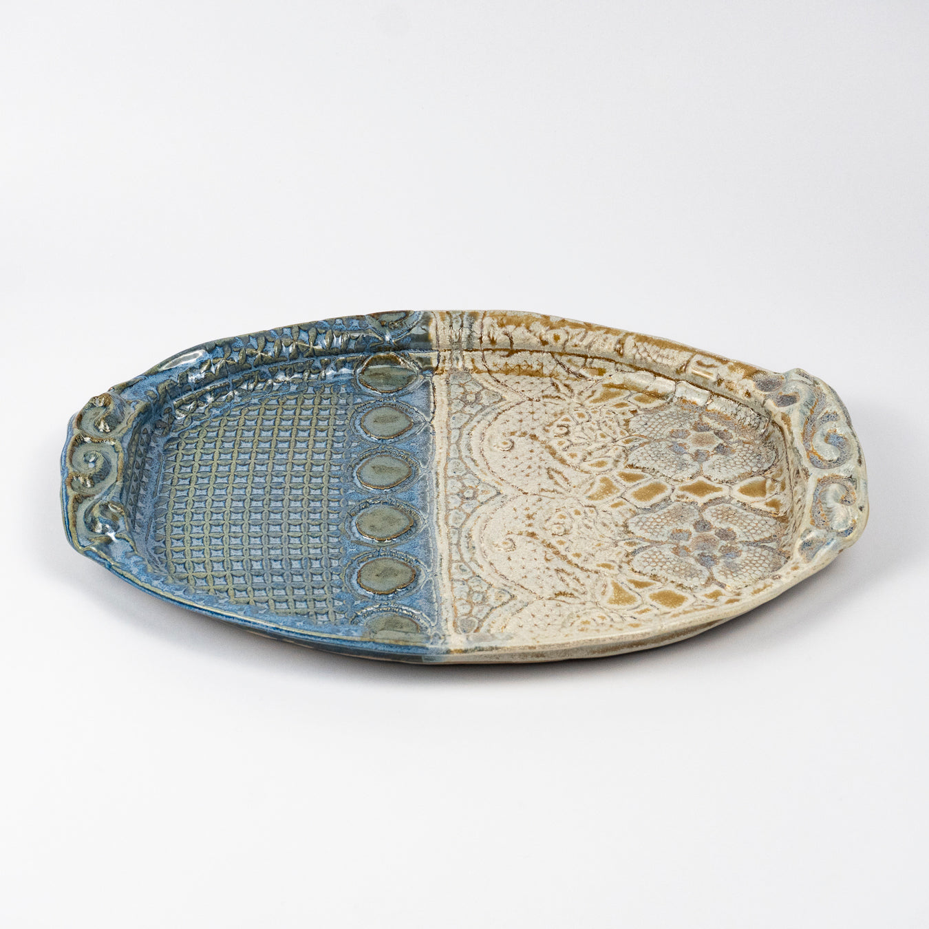 An oval ceramic serving tray with a split design, featuring a blue glazed grid pattern on one half and a beige glazed floral pattern on the other, set against a plain light gray background.
