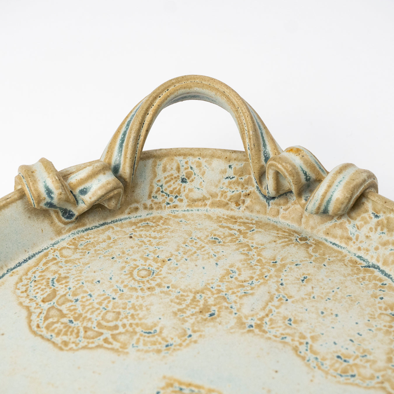 Close-up of a ceramic plate with a raised handle, featuring a textured beige and cream glaze with subtle blue-green speckles on a white background.