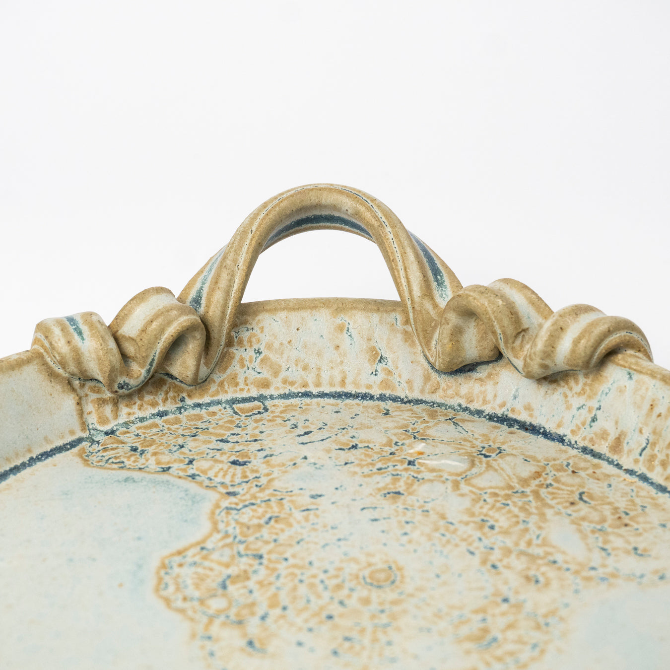 Close-up of a ceramic tray with a sculpted handle, featuring a speckled beige and white glaze with blue accents.