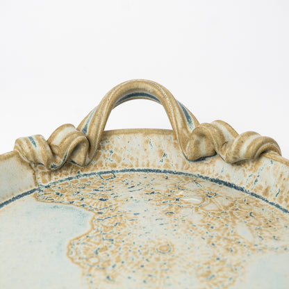 Close-up of a ceramic tray with a sculpted handle, featuring a speckled beige and white glaze with blue accents.