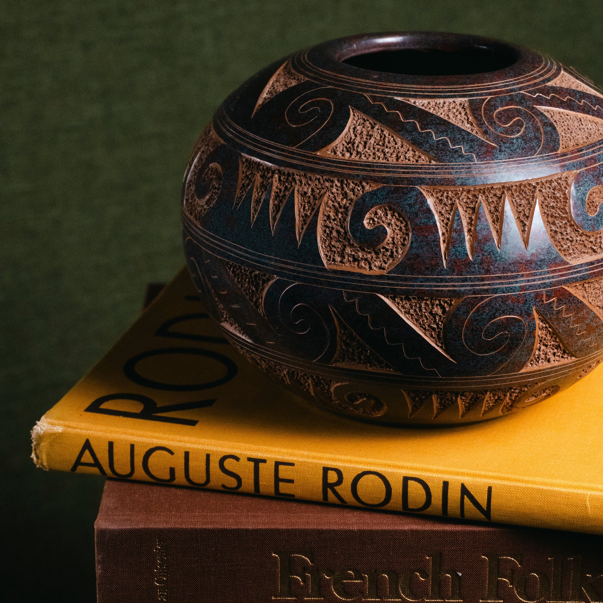 A decorative dark brown and black patterned ceramic vase sits on top of two books, with the top book showing the title 'Auguste Rodin' and the bottom book showing 'French Folk'