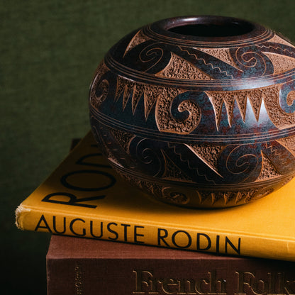 A decorative dark brown and black patterned ceramic vase sits on top of two books, with the top book showing the title 'Auguste Rodin' and the bottom book showing 'French Folk'