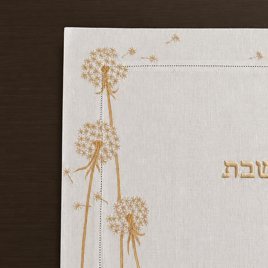 a close up detailed image of the Whispera cream linen challah cover with gold hand-embroidered dandelions along both sides, hemstitched border, and gold Shabbat lettering at center