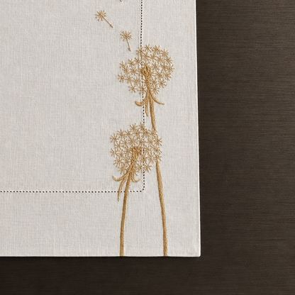 a detailed photo of the Whispera cream linen challah cover with gold hand-embroidered dandelions along both sides, hemstitched border, and gold Shabbat lettering at center