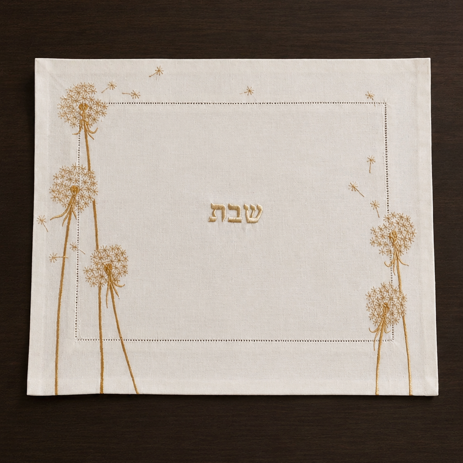 The Whispera cream linen challah cover with gold hand-embroidered dandelions along both sides, hemstitched border, and gold Shabbat lettering at center