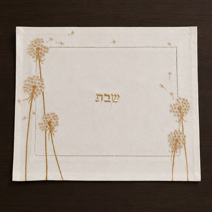 The Whispera cream linen challah cover with gold hand-embroidered dandelions along both sides, hemstitched border, and gold Shabbat lettering at center