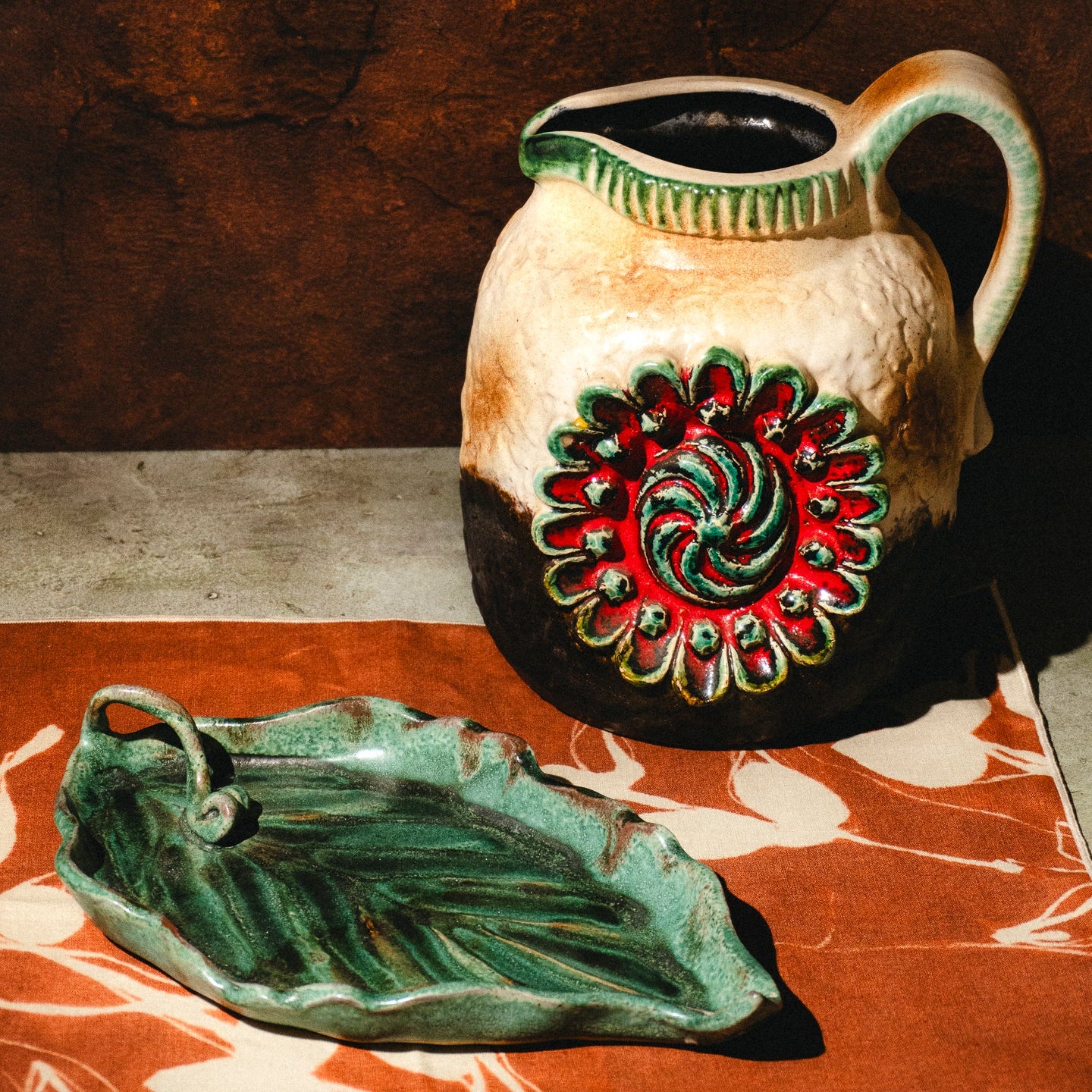 A rustic ceramic pitcher with a textured cream and dark brown glaze, decorated with a red and green floral motif on the front, shown alongside a matching leaf-shaped ceramic dish on a patterned surface.
