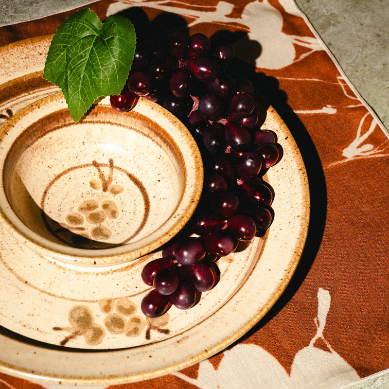 A ceramic bowl and matching plate with a floral pattern, styled with dark purple grapes and a green leaf on a brown and beige placemat.