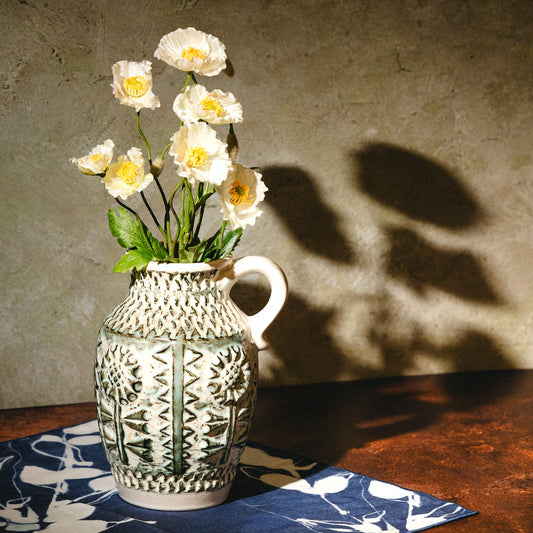 A white ceramic pitcher vase with green geometric patterns, filled with white flowers, set on a blue and white patterned cloth on a dark wooden table against a textured gray wall with leaf shadows.