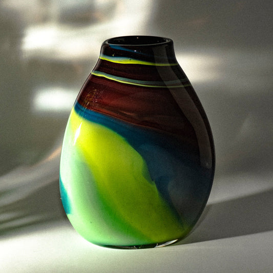 A hand-blown glass vase with an organic teardrop shape, featuring marbled swirls of dark brown, blue, neon green, and black, set against a softly lit gray background with dramatic light shadows.