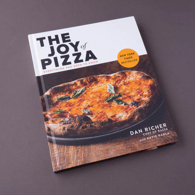 The Joy of Pizza cookbook by Dan Richer, featuring a white and dark brown cover with a photo of a freshly baked pizza, displayed on a dark gray surface.