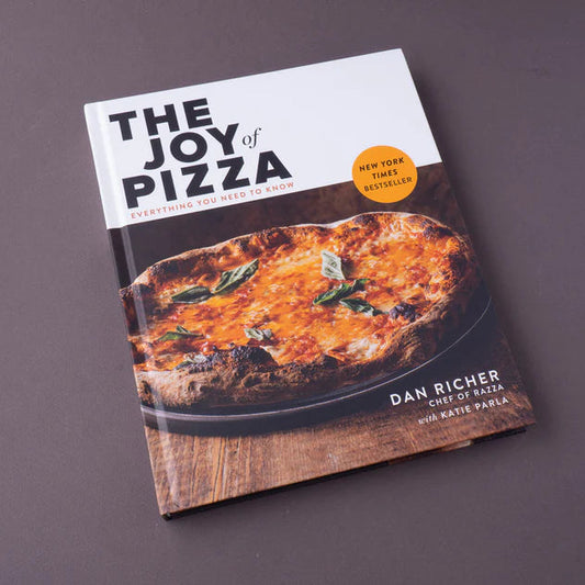 The Joy of Pizza cookbook by Dan Richer, featuring a white and dark brown cover with a photo of a freshly baked pizza, displayed on a dark gray surface.