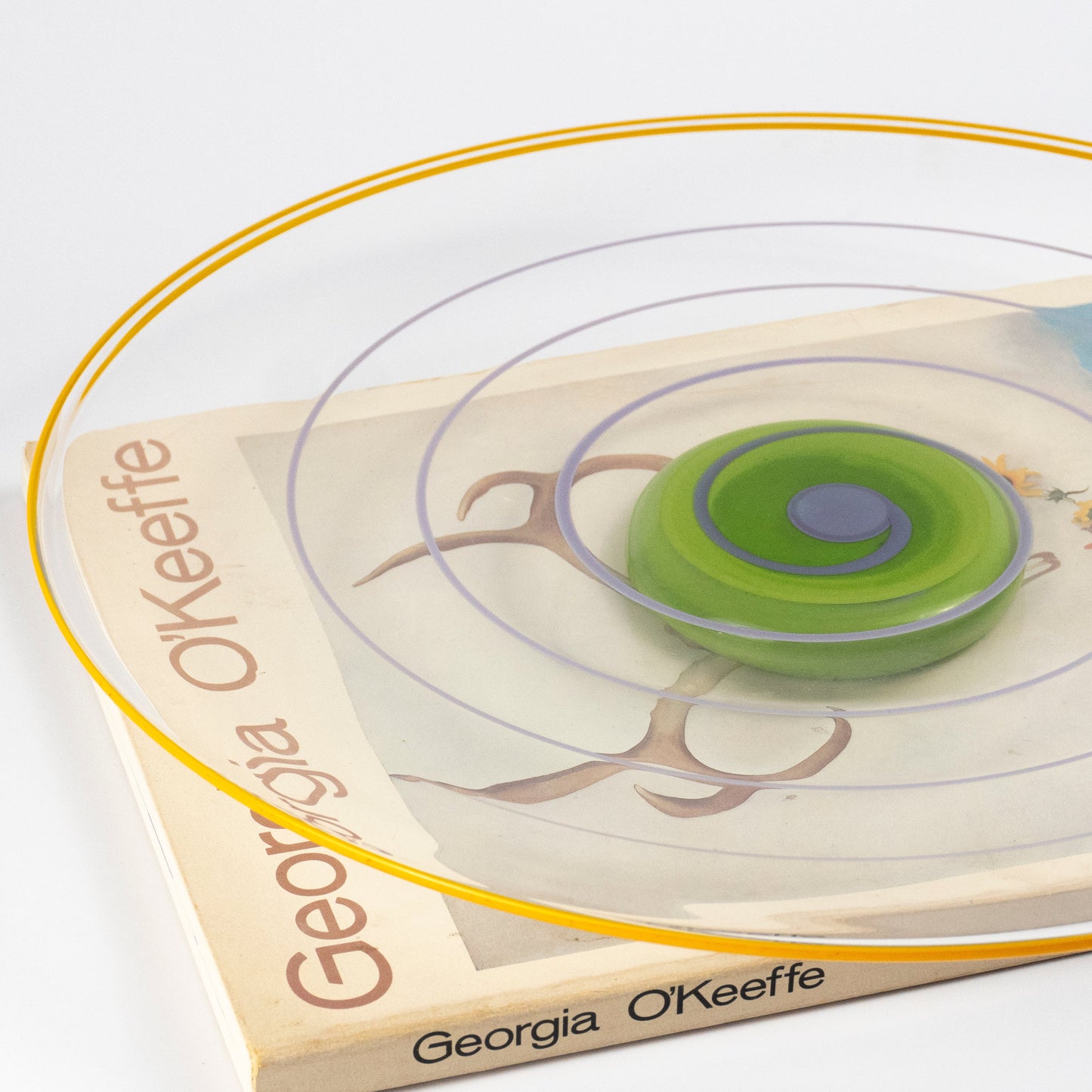 A side view of a clear circular plate with concentric rings in yellow, purple, and green, atop a 'Georgia O'Keefe' book 