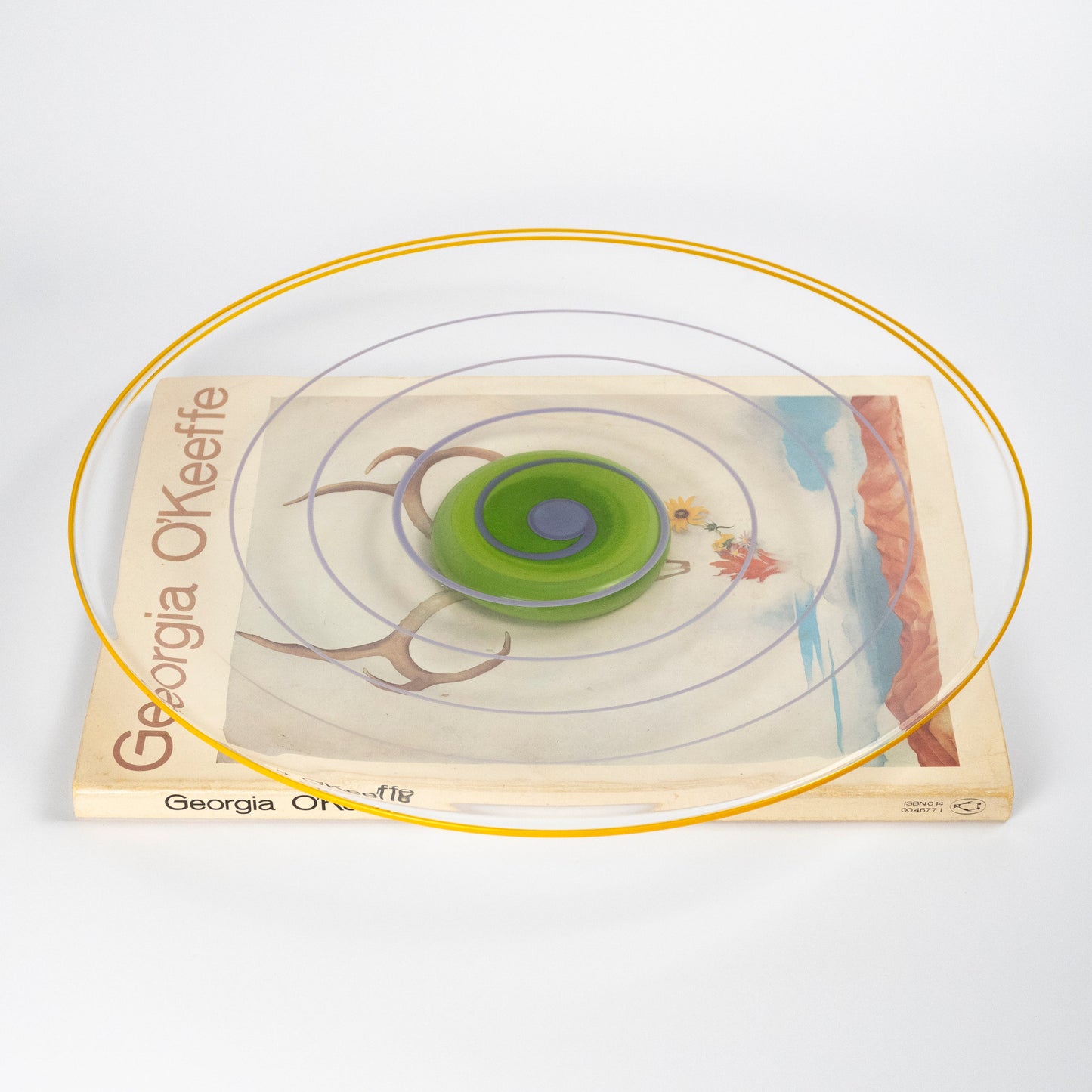 A top-down view of a clear circular plate with concentric rings in yellow, purple, and green, atop a 'Georgia O'Keefe' book 