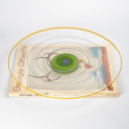 A top-down view of a clear circular plate with concentric rings in yellow, purple, and green, atop a 'Georgia O'Keefe' book 