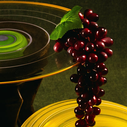 A close-up studio still life of a dark red grape bunch with a green leaf resting on a clear glass platter above a yellow plate, against a dark green background.