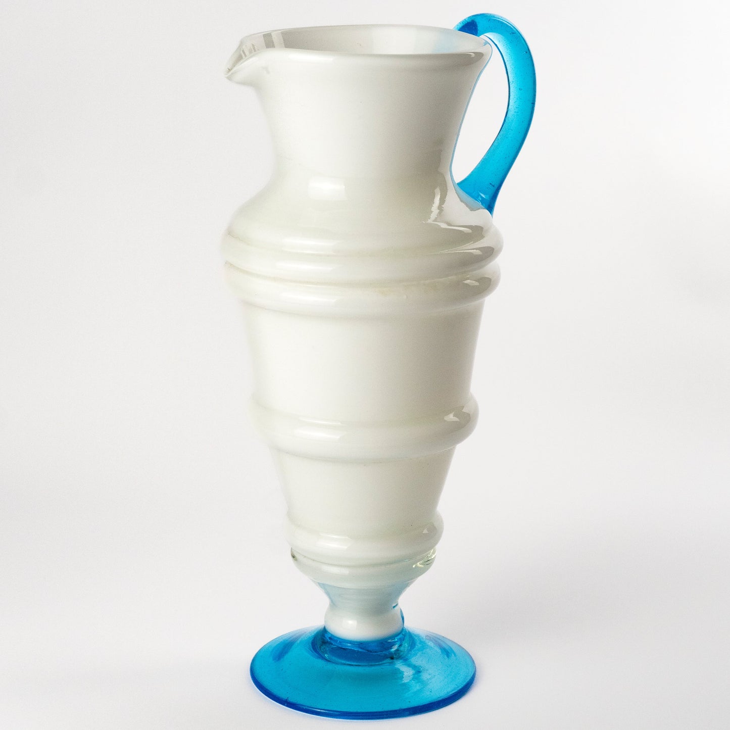 A white glass pitcher with a bright blue handle and matching blue footed base, photographed on a seamless white background.
