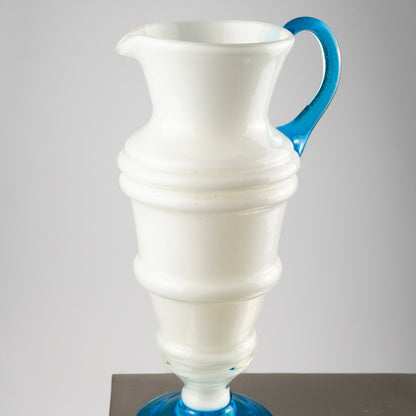 A white glass pitcher with a flared base, ribbed detailing, and a bright blue handle and matching blue circular foot, shown on a neutral gray background.