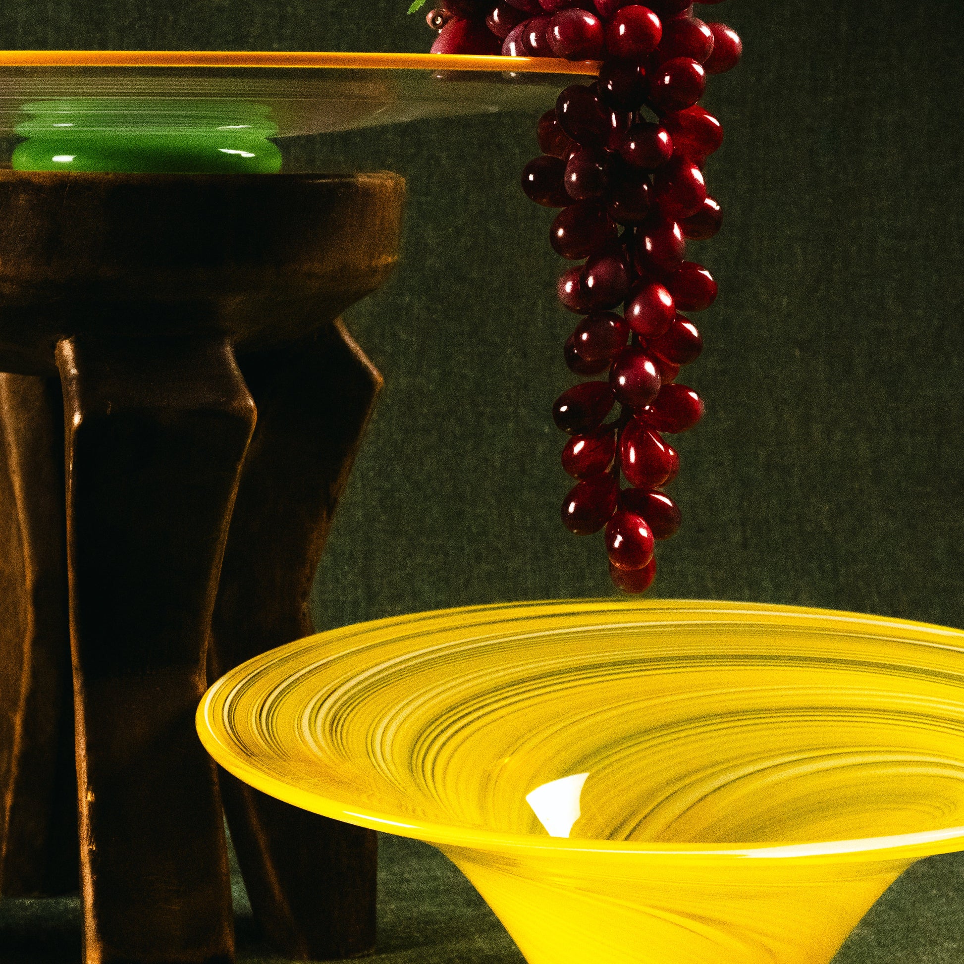 A yellow glass bowl with a wide rim in the foreground, a dark wooden stand holding a clear glass dish with green and red glass accents in the background, and a cluster of glossy dark red grapes hanging over the edge of the dish against a dark textured backdrop.