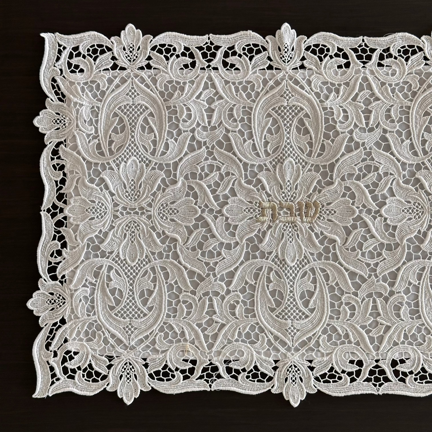 a white lace challah cover with an intricate floral and scroll pattern, laid flat on a dark brown wooden surface.