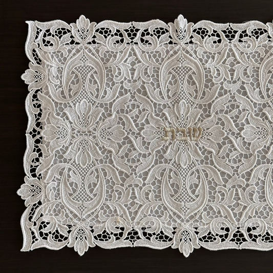 a white lace challah cover with an intricate floral and scroll pattern, laid flat on a dark brown wooden surface.