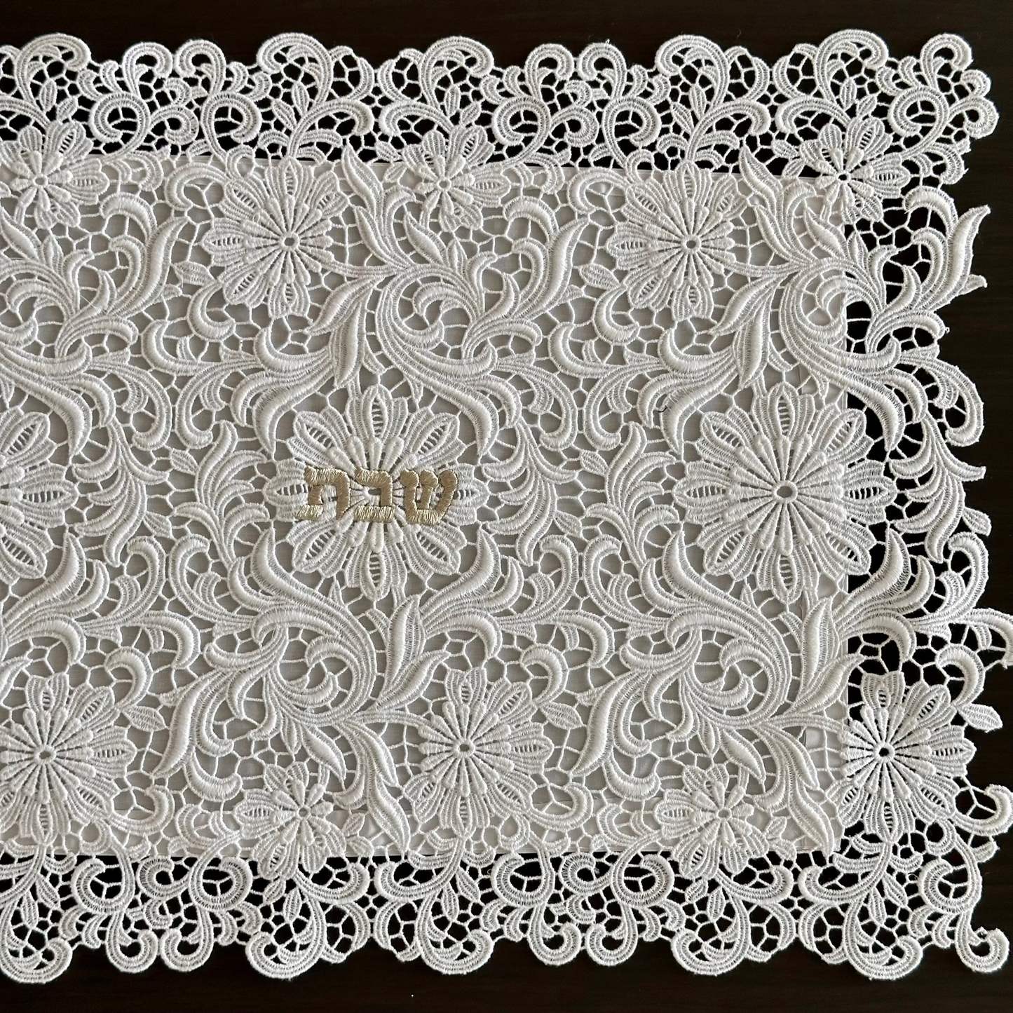White floral lace challah cover with intricate embroidered patterns and scalloped edges, laid flat on a dark brown wooden surface.