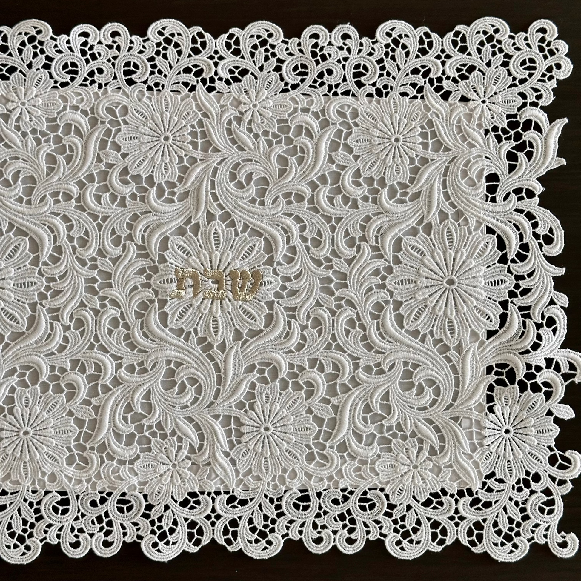White floral lace challah cover with intricate embroidered patterns and scalloped edges, laid flat on a dark brown wooden surface.