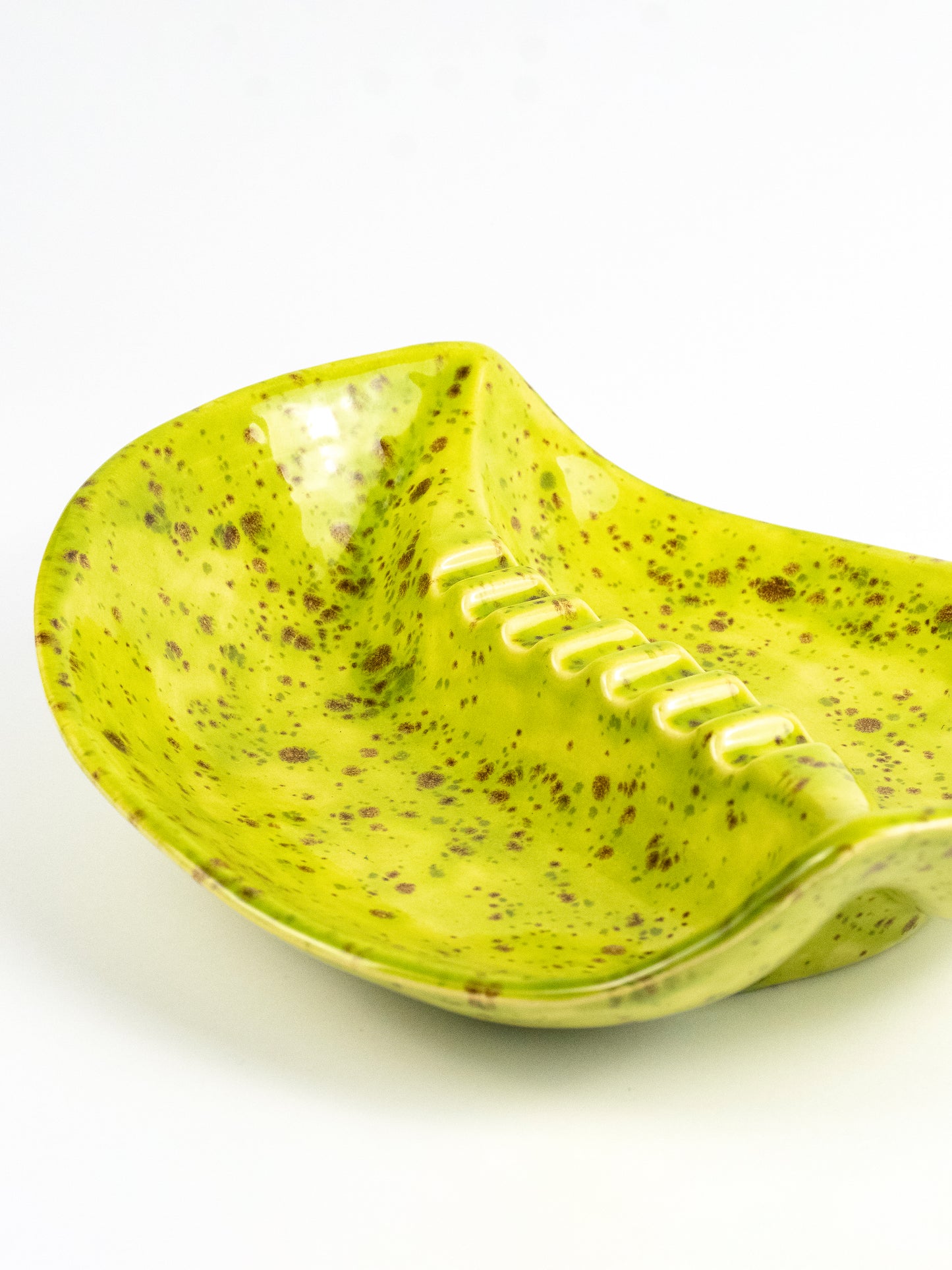 A ceramic bowl with a wavy, spoon-like form, featuring a bright lime green glaze speckled with brown and white spots, set against a plain white background.