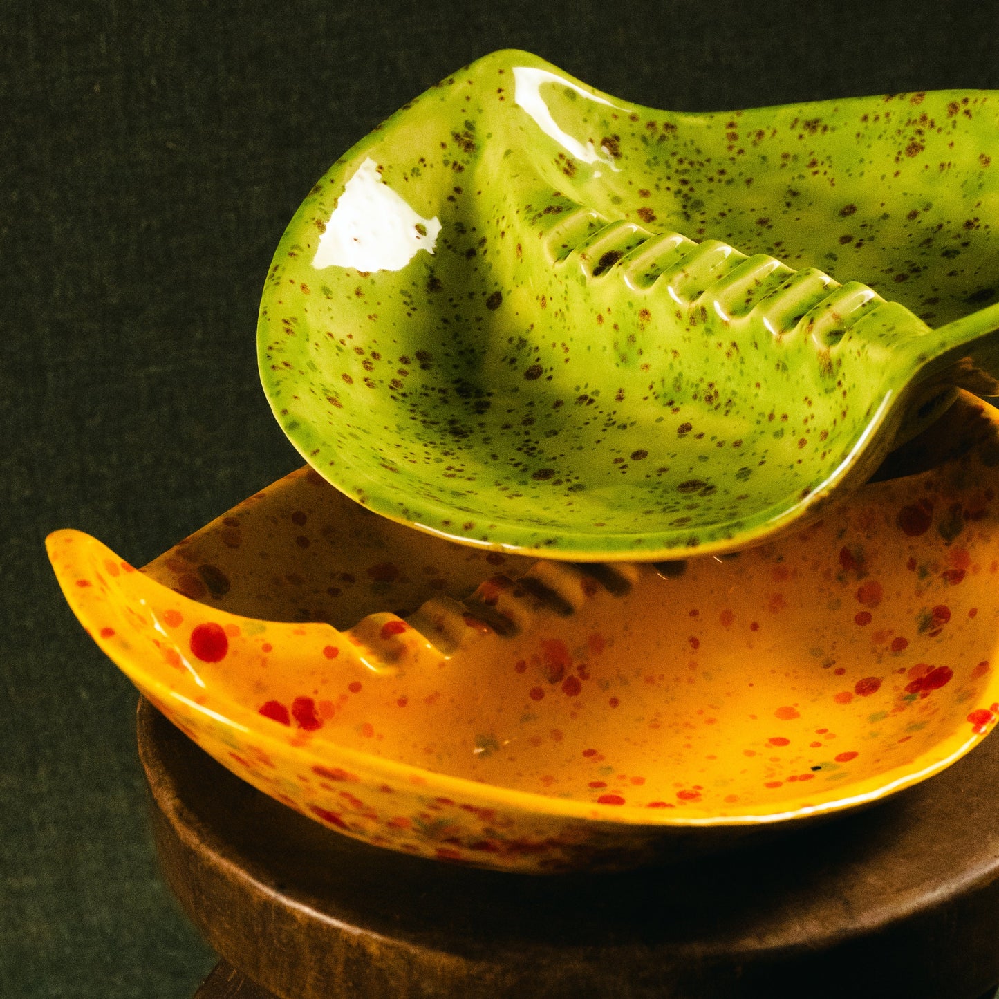 Two ceramic leaf-shaped bowls with speckled glazes, one green and one orange-yellow, displayed on a dark wooden stand against a dark background.
