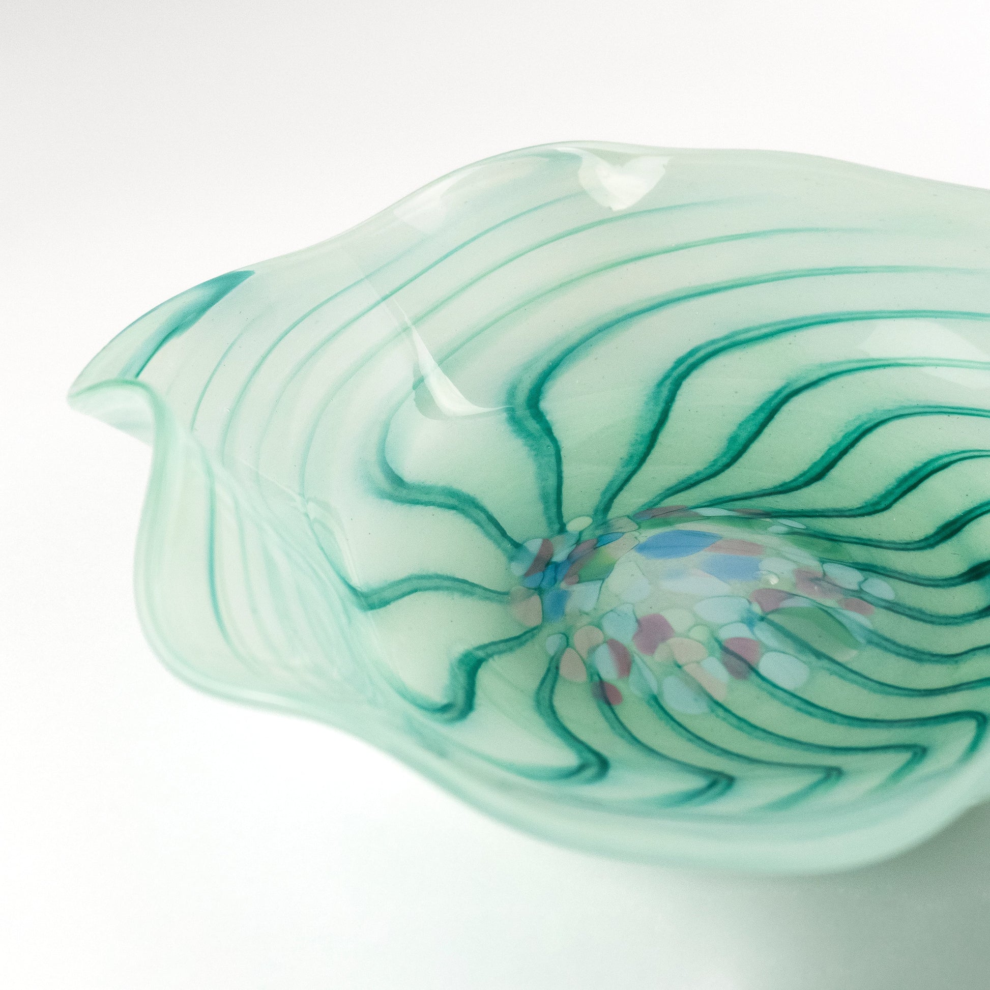 A hand-blown glass bowl with a wavy, organic shape, featuring a translucent pale mint green body with dark green wavy lines and a colorful bubble burst near the center, set against a clean white background.