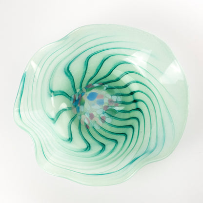 A hand-blown glass bowl with a wavy, scalloped rim, featuring a light mint green base with concentric teal-green swirls and a small multicolored center detail, set against a plain white background.