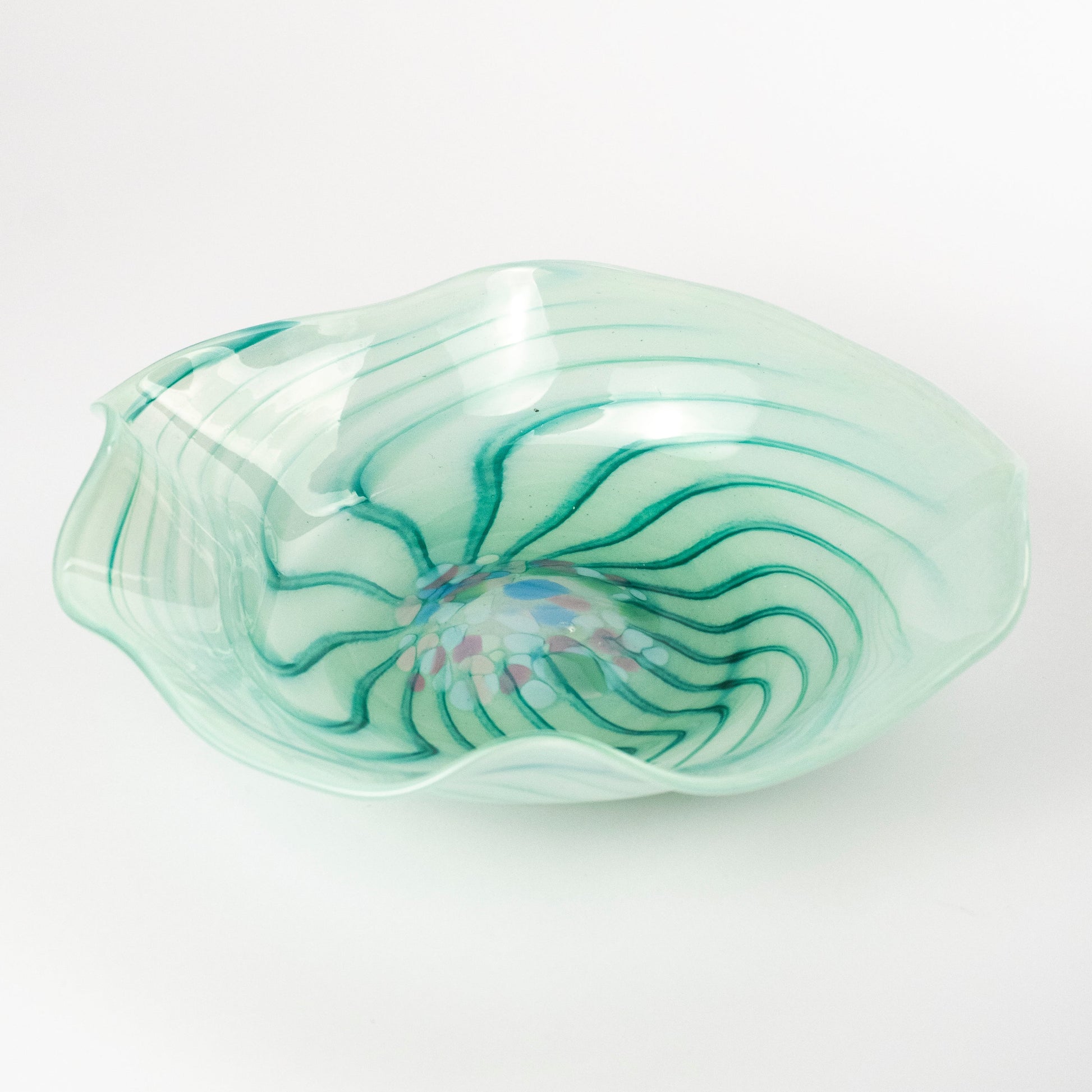 A hand-blown glass bowl with a wavy, organic shape, featuring a light mint green base with dark green swirling patterns and a small iridescent blue and pink center, set against a plain white background.
