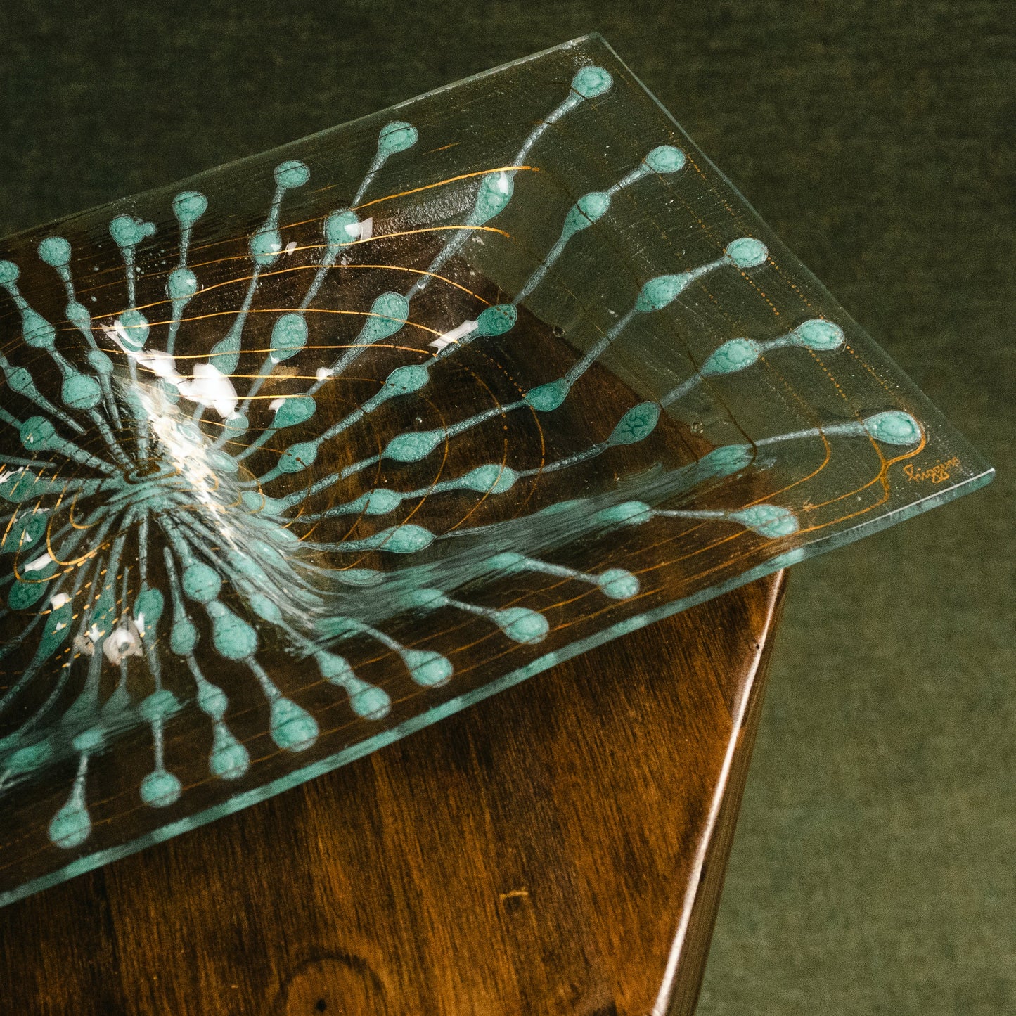 A clear rectangular glass plate with a turquoise and gold dendritic pattern, resting on a dark wooden surface against a dark textured background.