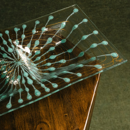 A clear rectangular glass plate with a turquoise and gold dendritic pattern, resting on a dark wooden surface against a dark textured background.