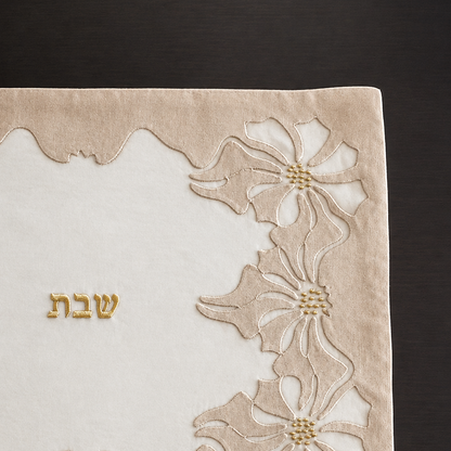 A rectangular beige and white linen and organza embroidered challah cover with floral motifs in the corners and the Hebrew word 'Shabbat' in gold in the center, laid flat on a dark wood background.