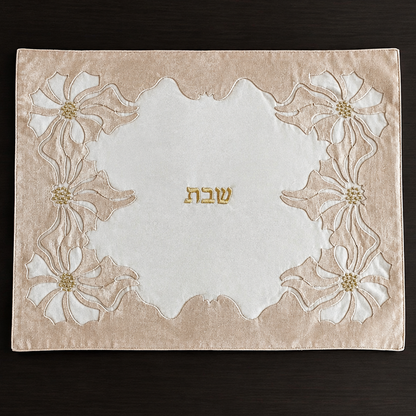 A rectangular beige and white linen and organza embroidered challah cover with floral motifs in the corners and the Hebrew word 'Shabbat' in gold in the center, laid flat on a dark wood background.