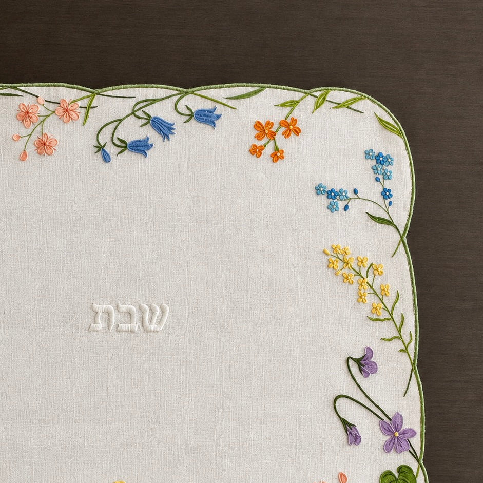 A hand embroidered Shabbat challah cover with a scalloped border featuring colorful wildflowers and green leaves on a cream linen background, with the Hebrew word 'Shabbat' embroidered in the center, displayed on a dark brown wooden surface.