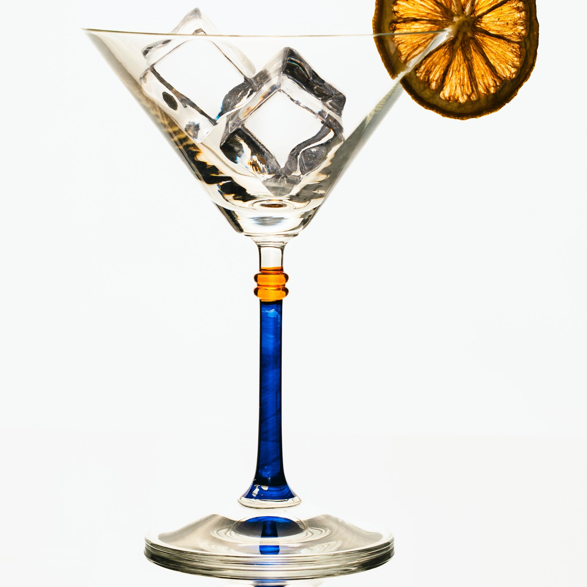 A martini glass with a blue stem and an orange slice garnish, filled with ice cubes, set against a clean white background.
