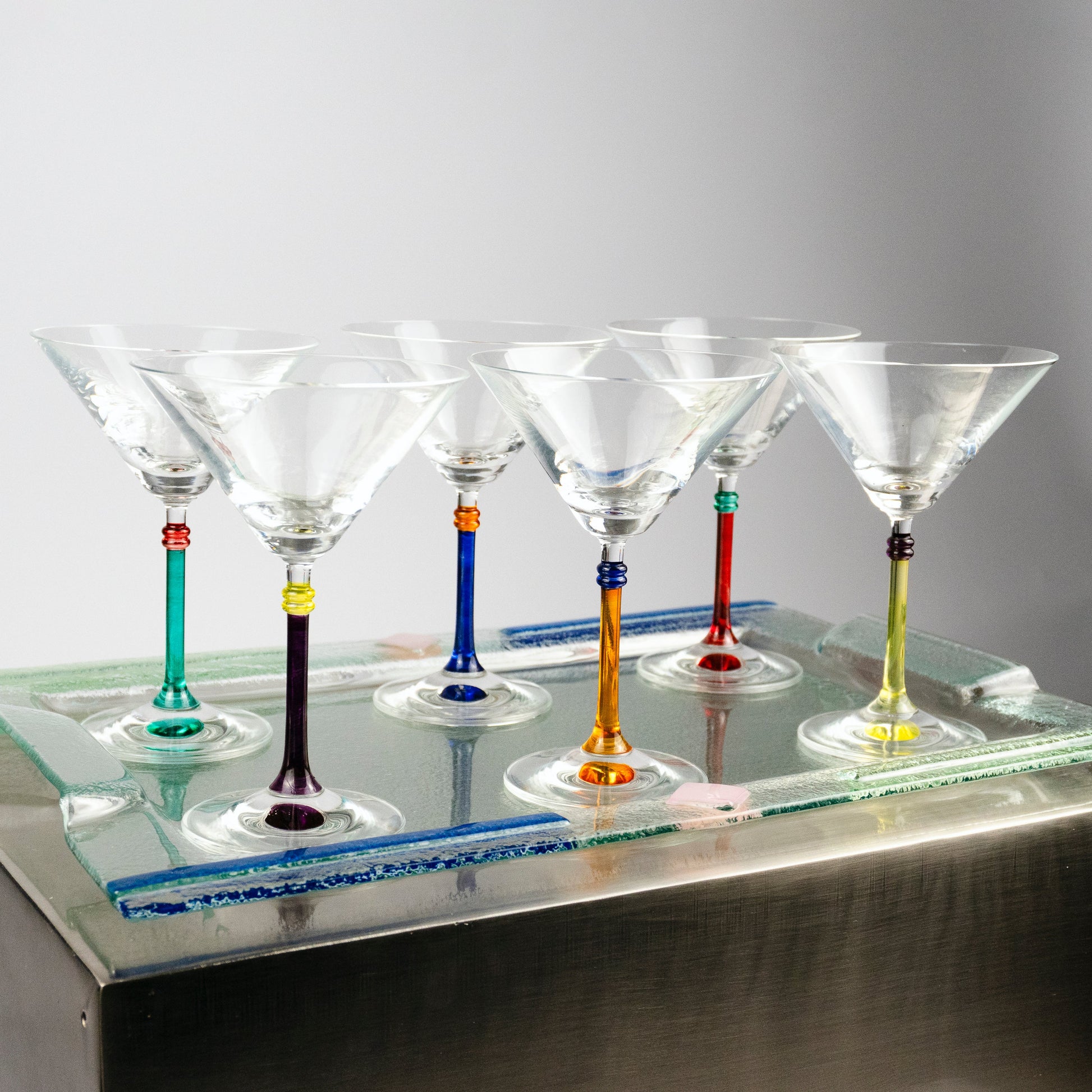 Six clear martini glasses with colorful stems in teal, purple, blue, orange, red, and yellow, arranged on a mirrored glass tray over a metallic surface against a light gray background.
