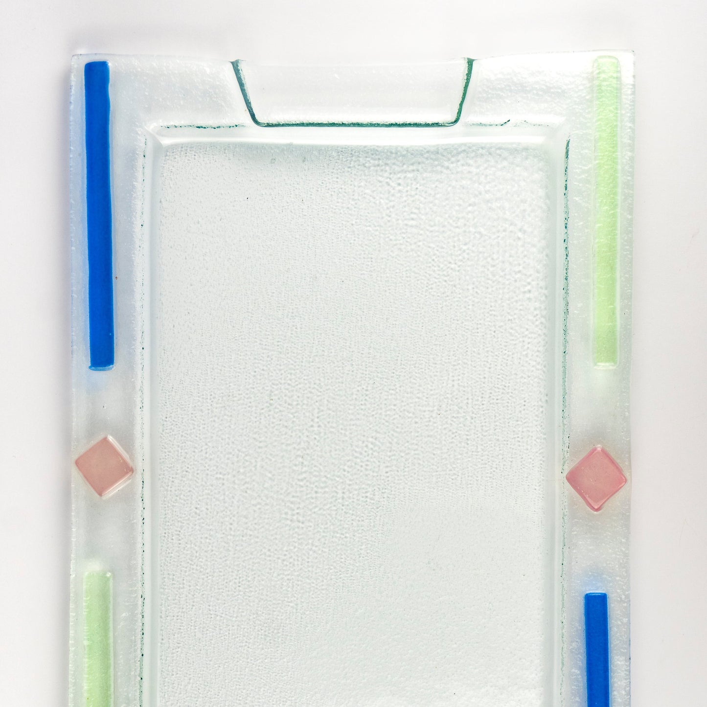 A square clear glass tray with a textured frosted center, featuring blue and light green geometric accents and small pink diamond shapes on a white background.