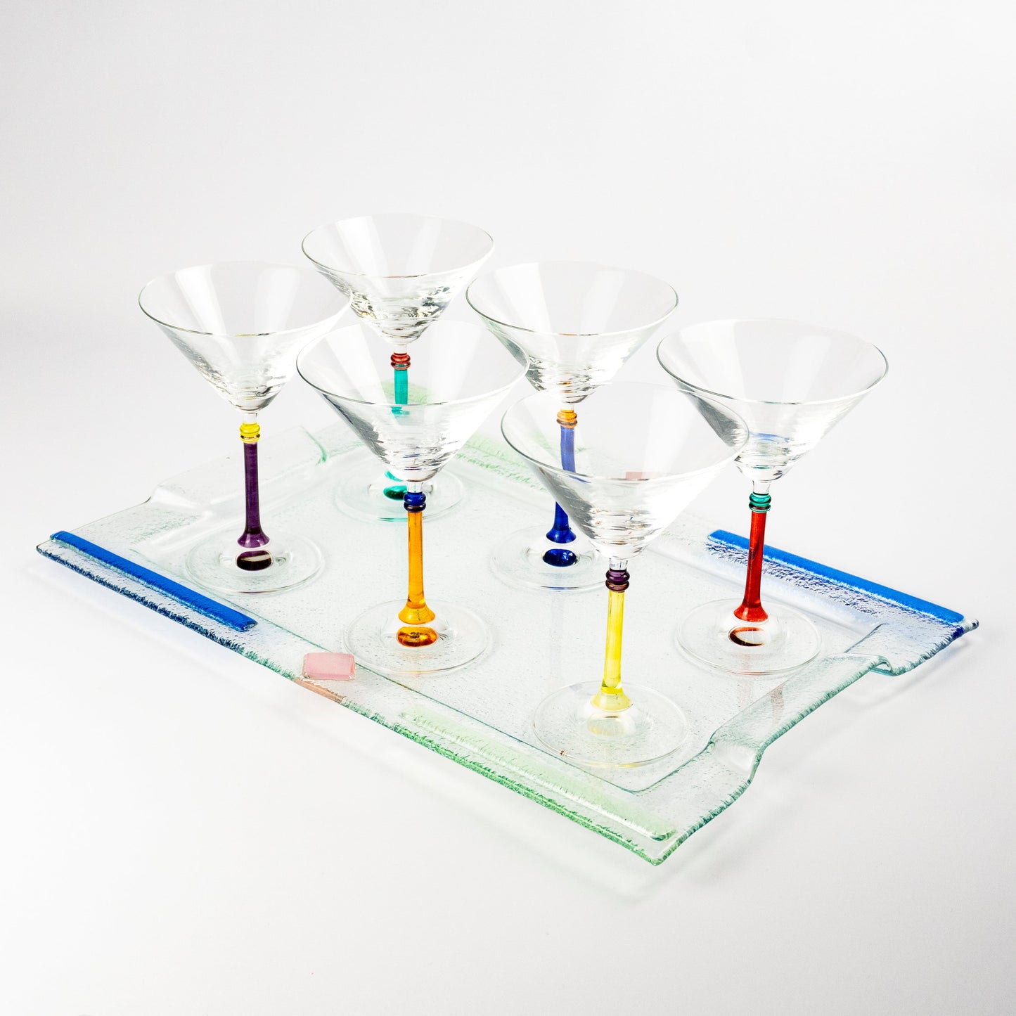 Six clear martini glasses with colorful glass stems in purple, green, orange, blue, and red, arranged on a clear rectangular glass tray against a white background.