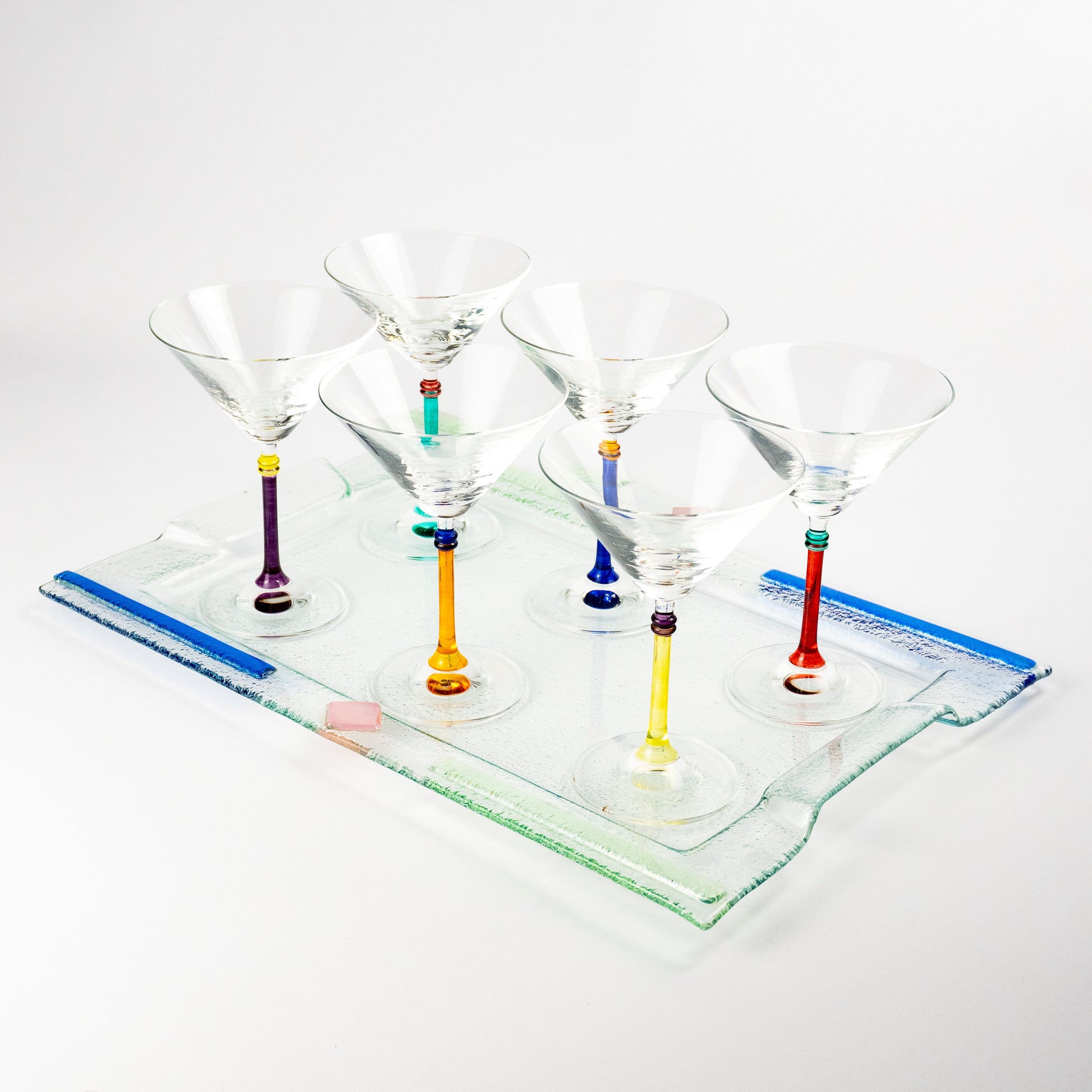 Six clear martini glasses with colorful glass stems in purple, green, orange, blue, and red, arranged on a clear rectangular glass tray against a white background.