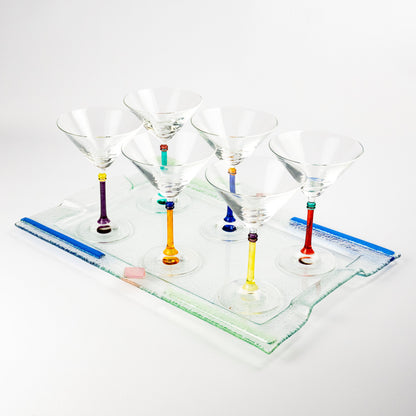 Six clear martini glasses with colorful glass stems in purple, green, orange, blue, and red, arranged on a clear rectangular glass tray against a white background.