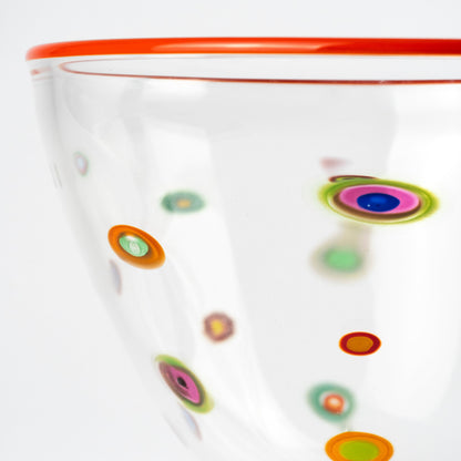 A close-up of a clear glass bowl with a vibrant red rim and base, decorated with colorful hand-blown glass dots known as Murrine in green, pink, purple, and orange.
