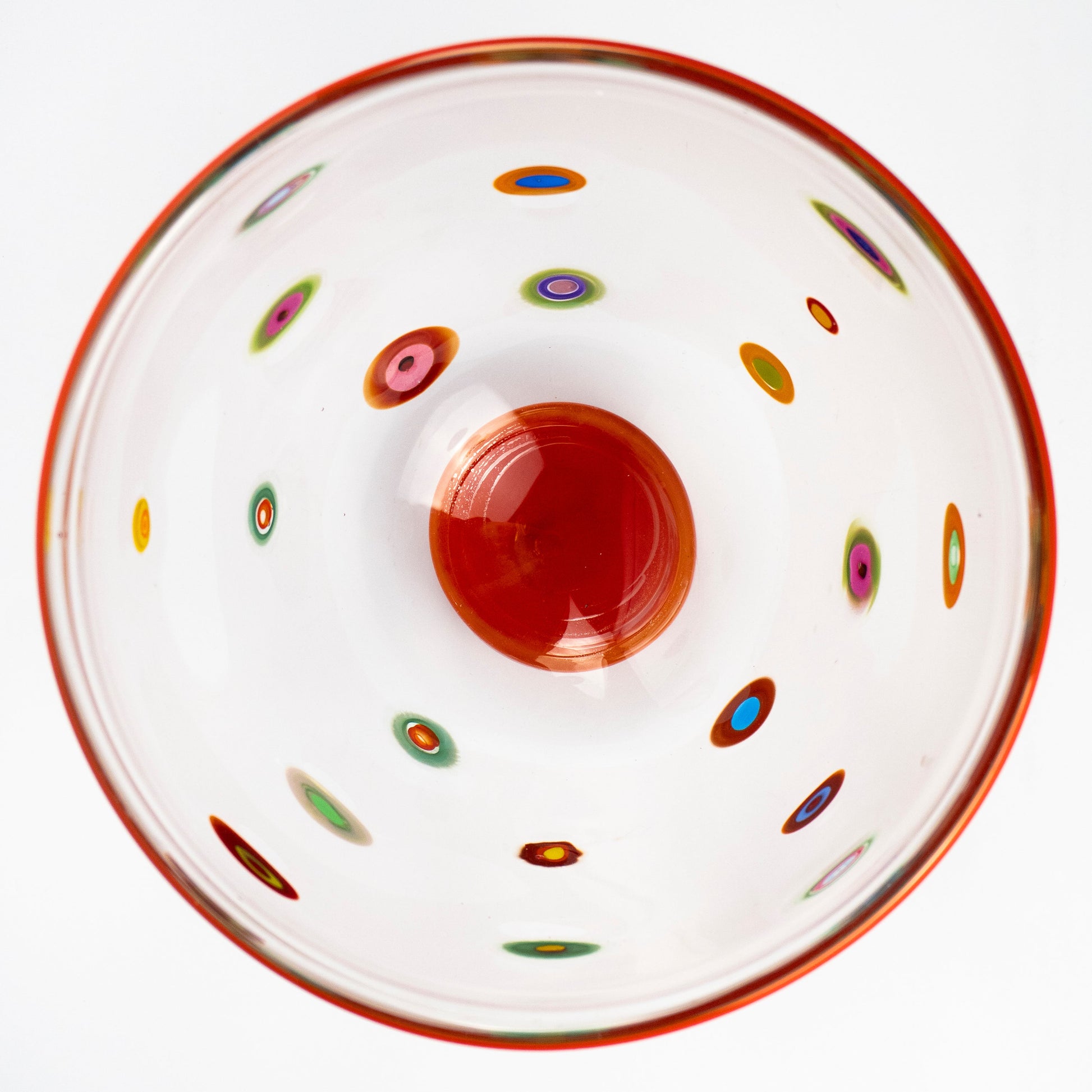 A top-down view of a clear glass bowl with a vibrant orange-red rim, a matching orange-red center, and multiple small multicolored concentric dot patterns scattered across the interior, set against a plain white background.